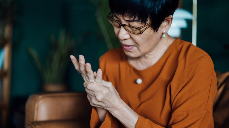 Senior Asian woman rubbing her hands in discomfort, suffering from arthritis in her hand while sitting on sofa at home. Elderly and health issues concept